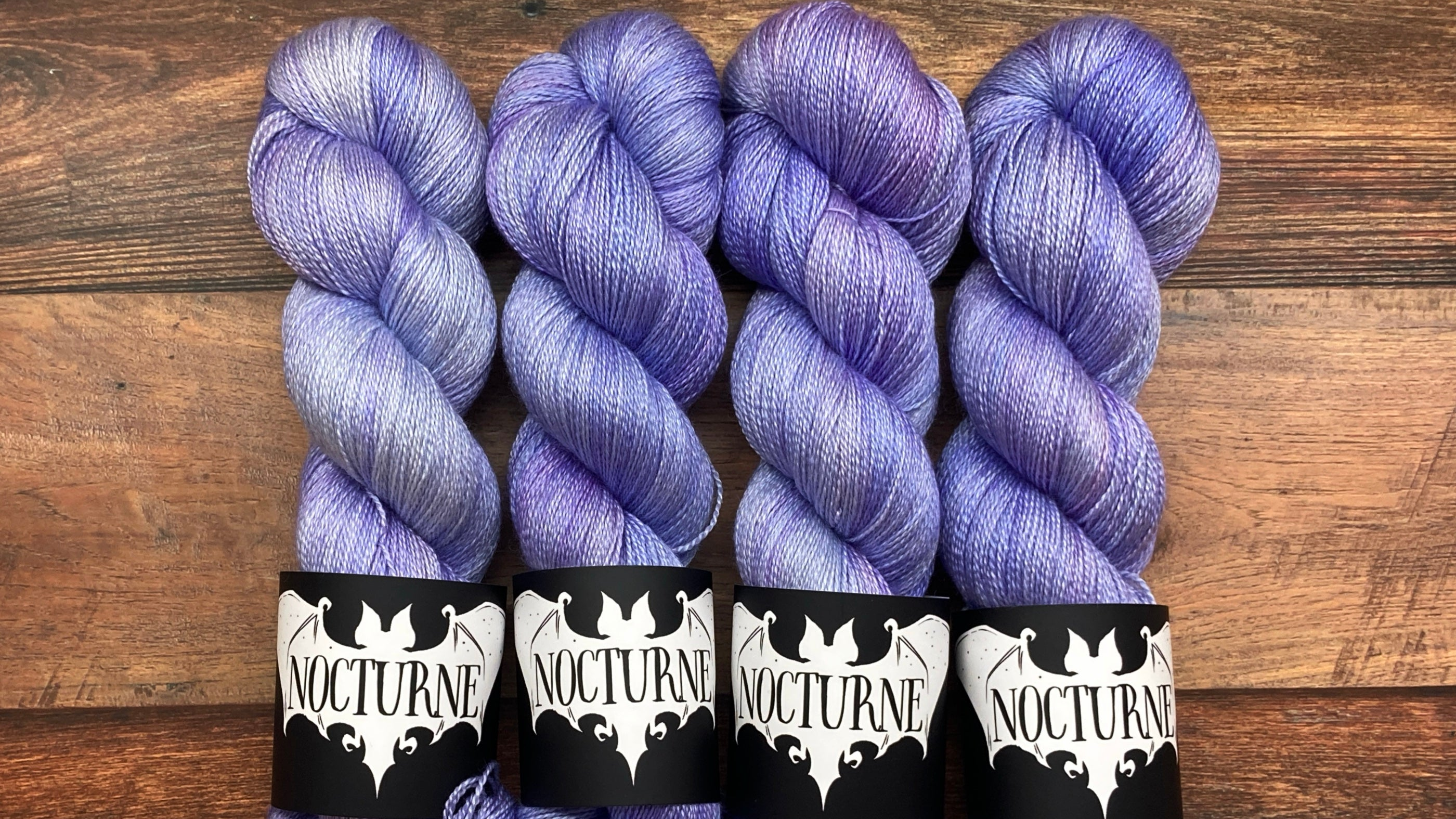 Four skeins of variegated purple and blue yarn with black and white 'Nocturne' paper bands, arranged side-by-side on a rustic wooden background.